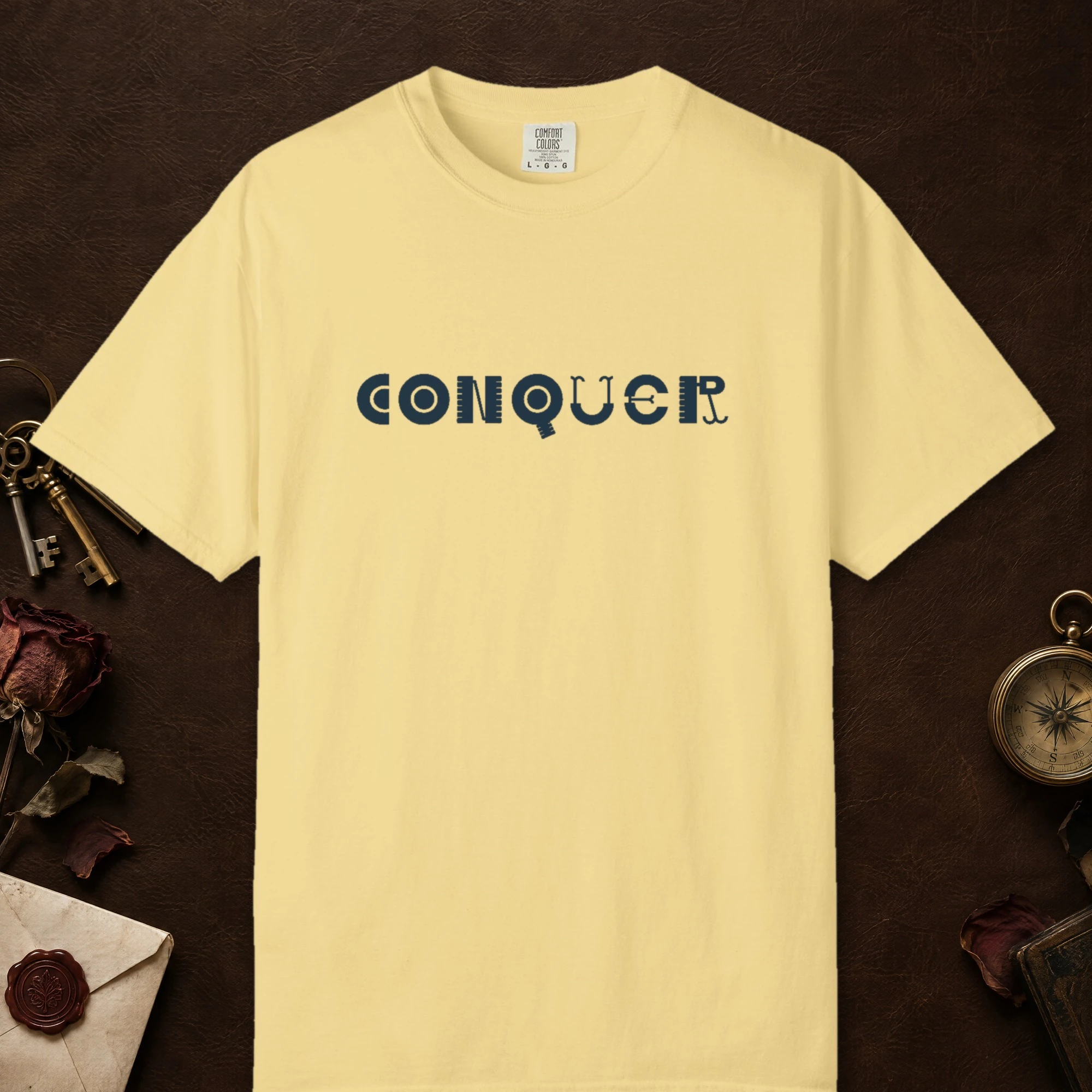 Conquer design on a Butter Comfort Colors 1717 tee, laid flat