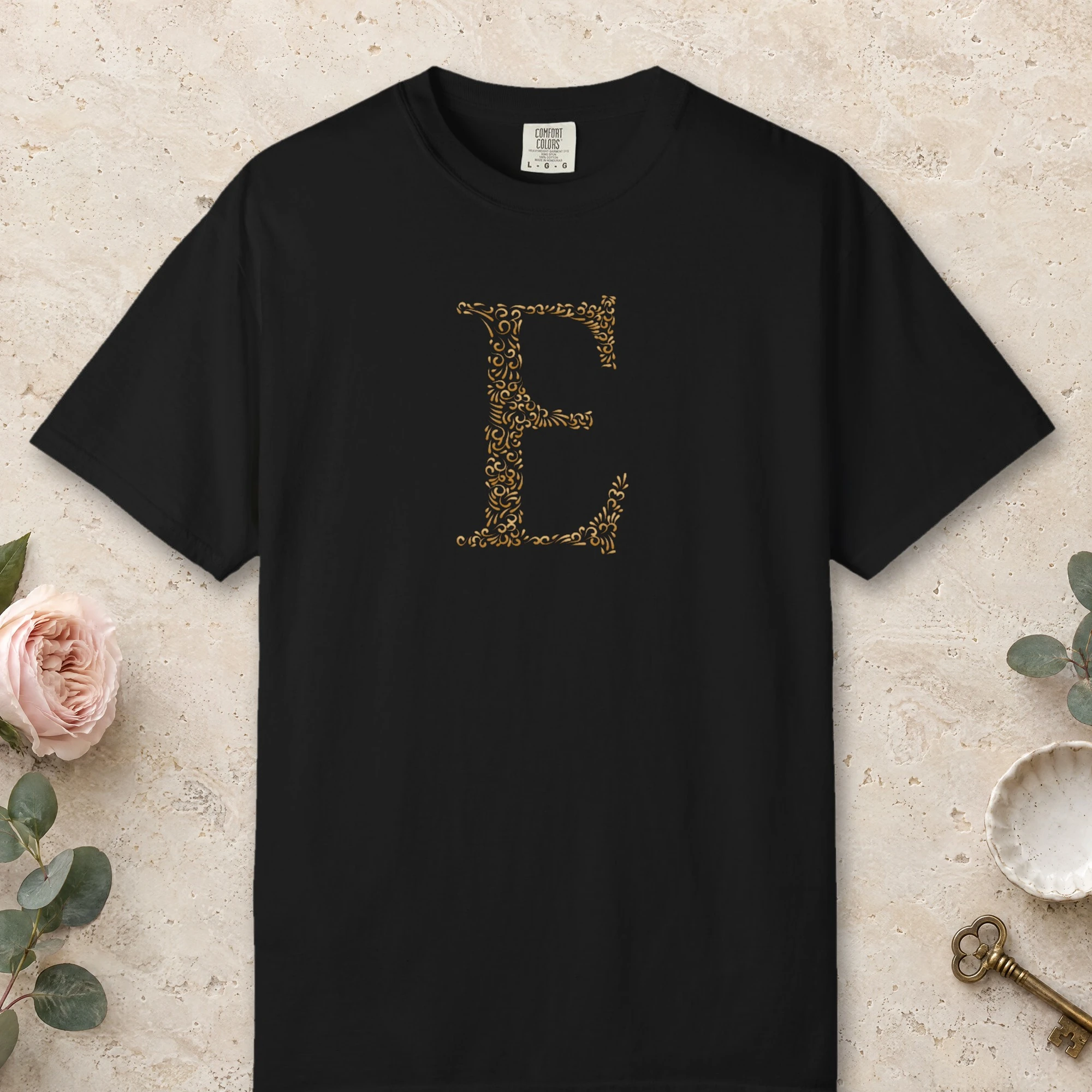 Initial E design on a Black Comfort Colors 1717 tee, laid flat