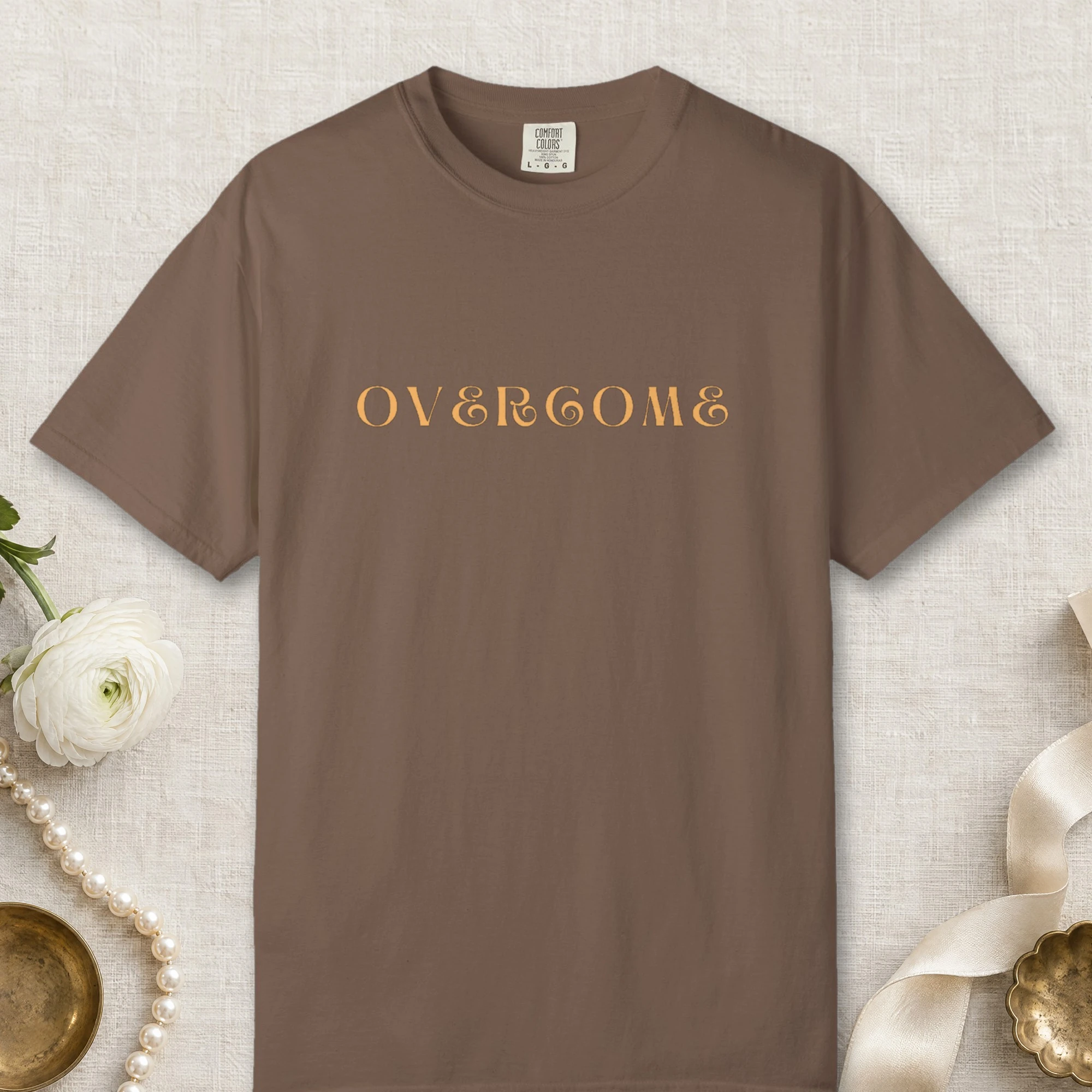 Overcome design on a Espresso Comfort Colors 1717 tee, laid flat