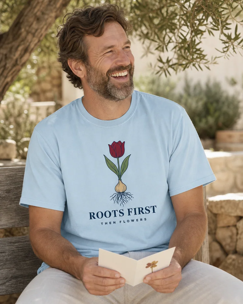 A father laughing over a handmade card on a garden bench on Father's Day (the father in the Roots First Tee in Chambray)