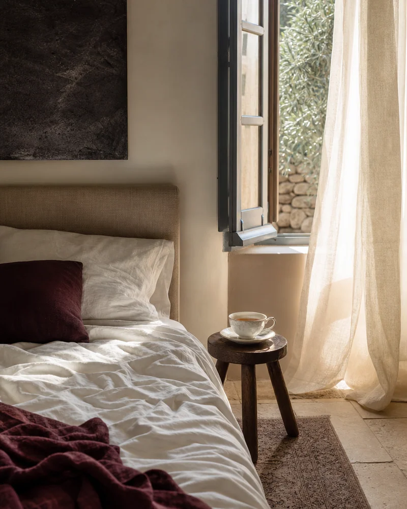 A bed of rumpled white linen in morning light with a cup of tea on a stool by an open window