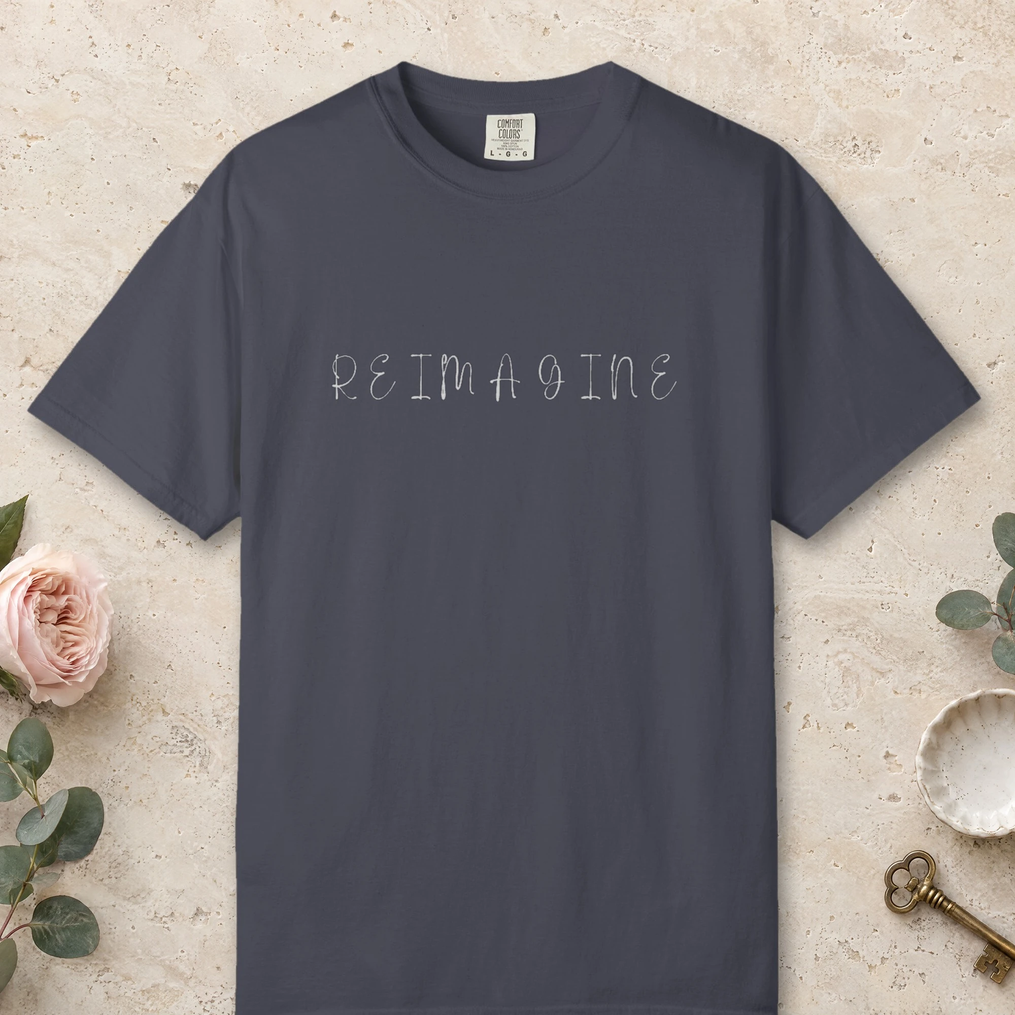 Reimagine Handwritten Capitals design on a Navy Comfort Colors 1717 tee, laid flat